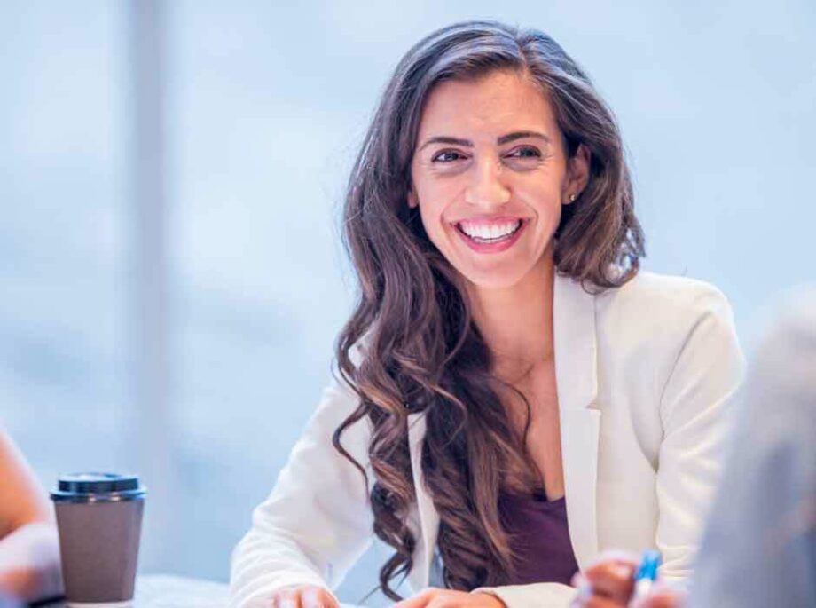 A smiling woman with long wavy brown hair sits at a table engaged in conversation