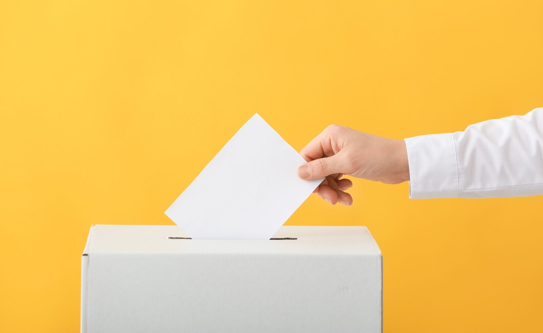 Hand placing piece of paper in voting box on yellow background