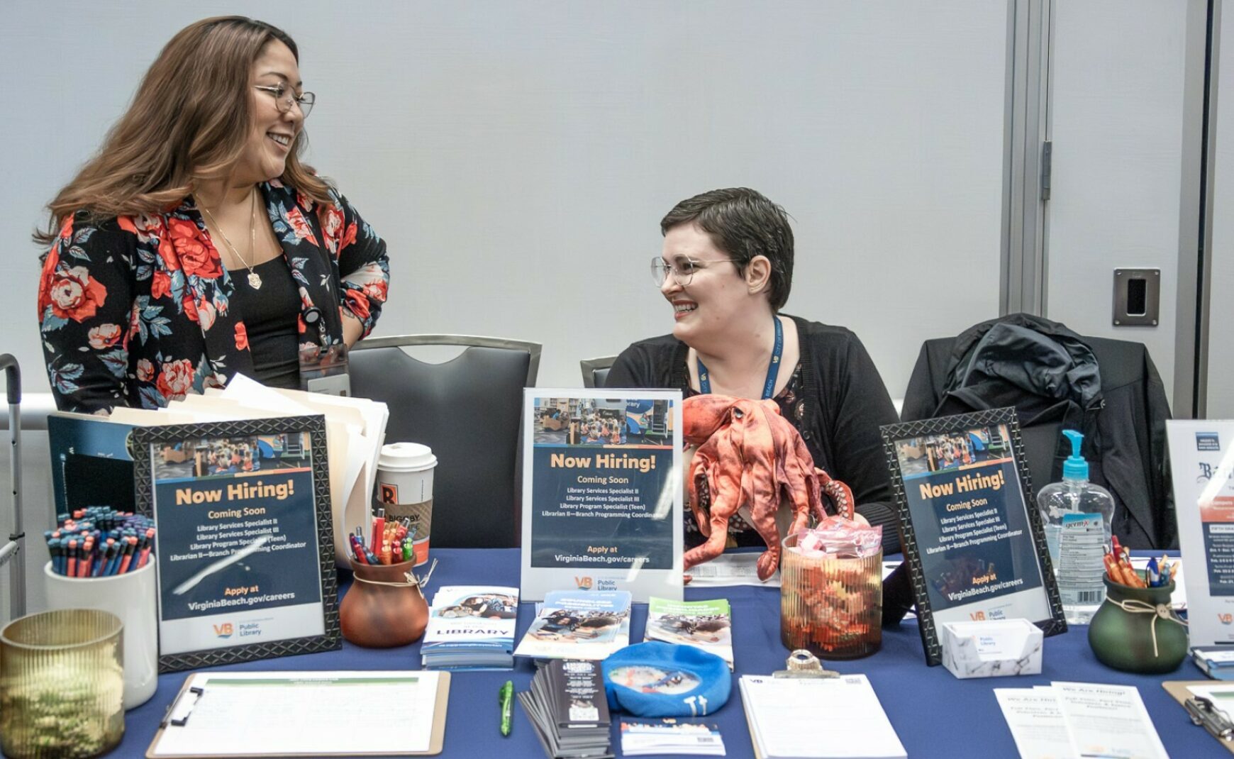 Now hiring signs at table at Virginia Beach Career Fair and two ladies smiling