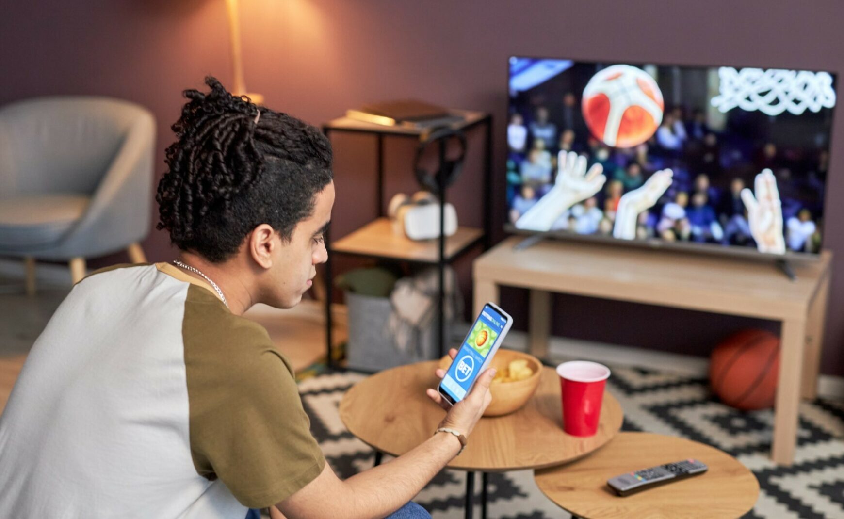 Person sitting on a couch engaged with a smartphone displaying a sports bet app while basketball game is on tv