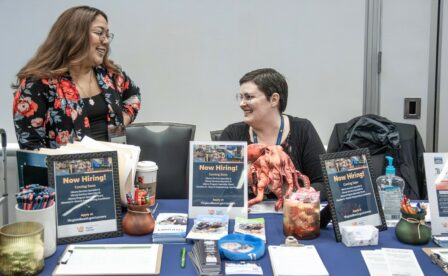 Now hiring signs at table at Virginia Beach Career Fair and two ladies smiling