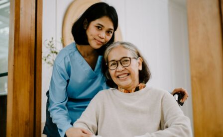 Caregiver standing behind elderly woman who is smiling and sitting in wheelchair
