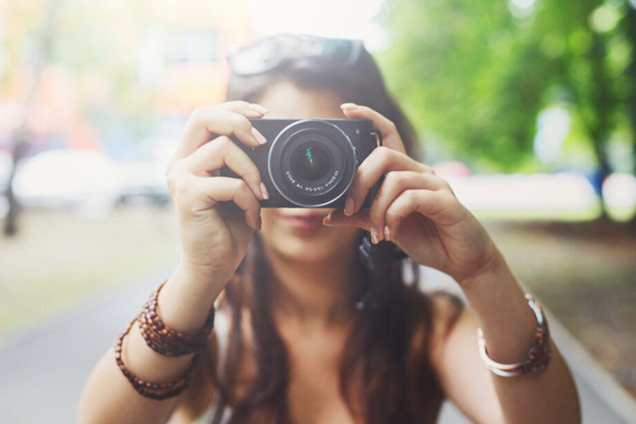 A woman wearing sunglasses holds up a camera while walking in a park.
